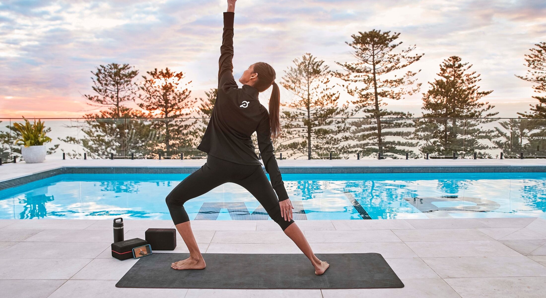Woman in all black leggings and jacket doing yoga on a mat in front of Manly Pacific pool and view of Manly Beach.